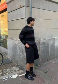 Maglione a righe nere, pantaloncini neri larghi e scarpe nere lucide. Superfici strutturate, tonalità scure variegate. In piedi contro un muro di pietra.
