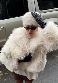 Witte mantel van nepbont met gestructureerde mouwen, zwarte leren handschoenen, stijlvolle zonnebril en een lichte bontmuts. Op de achtergrond een zilveren voertuig.