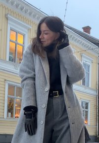 Femme en manteau gris, gants noirs et pantalon gris, debout à l'extérieur devant un bâtiment en bois jaune aux fenêtres éclairées en plein jour.
