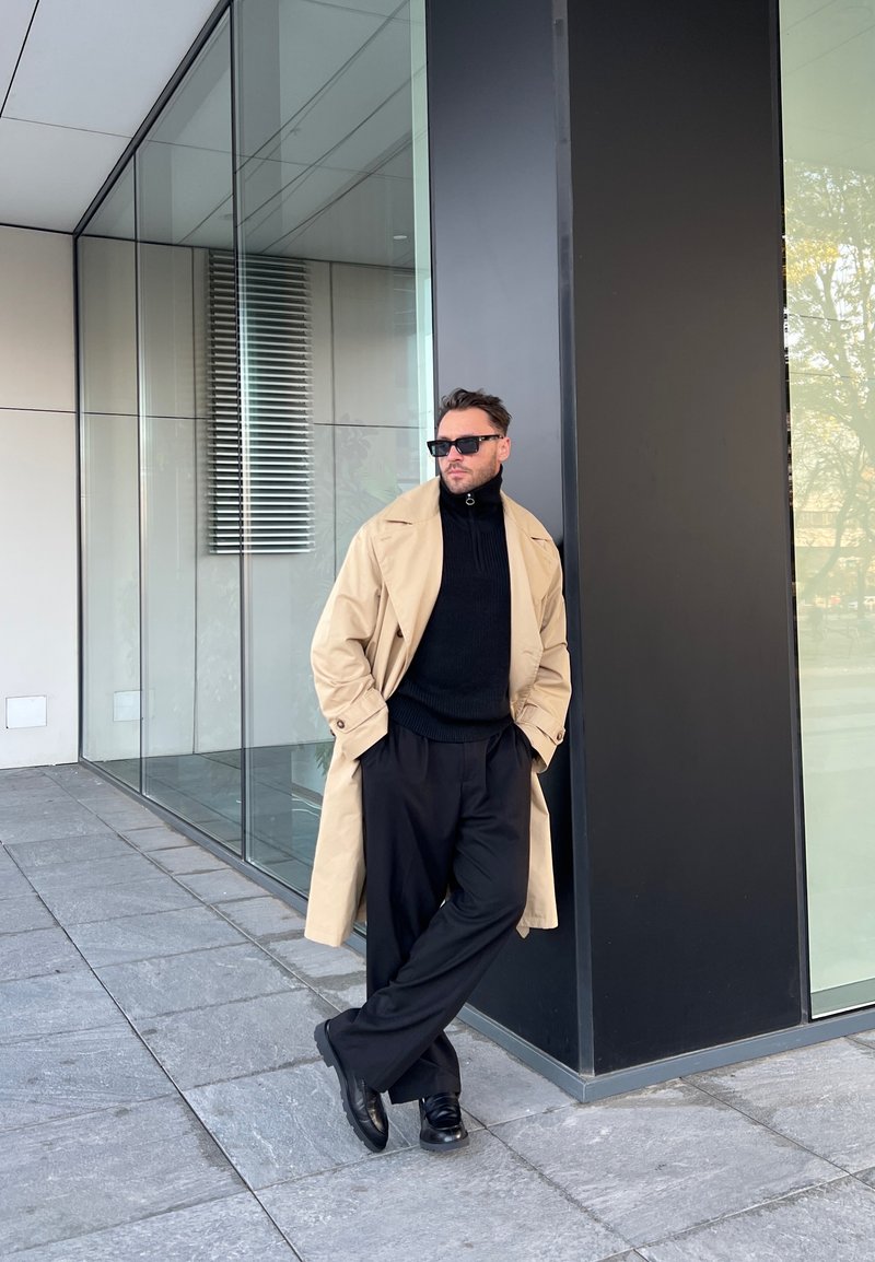 Κατα trench coat πάνω από ένα μαύρο ζιβάγκο, συνδυασμένο με μαύρα παντελόνια και παπούτσια. Στηρίζεται σε έναν γυάλινο τοίχο, με φόντο ένα σύγχρονο κτίριο.