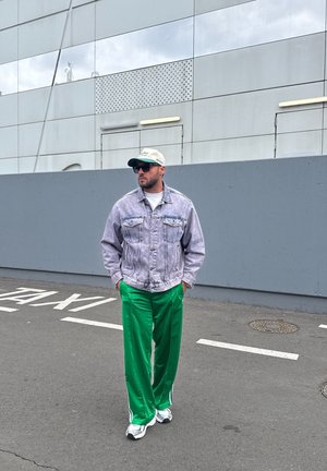 Chaqueta de mezclilla clara, camiseta blanca, pantalones deportivos verdes, zapatillas blancas y gorra de béisbol. De pie sobre el pavimento con un fondo de pared gris.