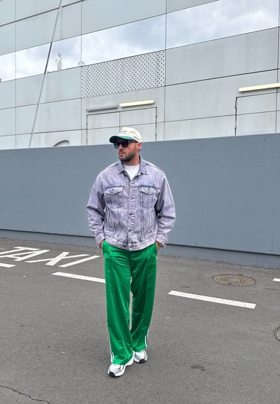 Chaqueta de mezclilla clara, camiseta blanca, pantalones deportivos verdes, zapatillas blancas y gorra de béisbol. De pie sobre el pavimento con un fondo de pared gris.
