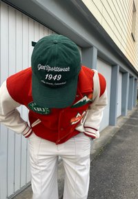 Casaco varsity de lã vermelho com mangas brancas, apresentando emblemas verdes e brancos. Boné verde com texto branco. Calças brancas.