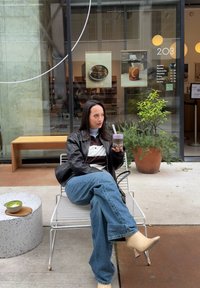 Una persona seduta tiene in mano una bevanda viola, indossando una giacca di pelle nera sopra una camicia a righe, jeans blu e stivali beige con tacco.