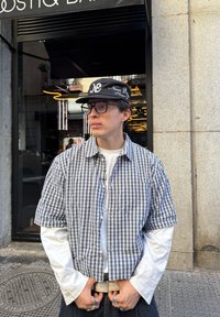 Koszula w kratę z krótkim rękawem w kolorze granatowym i białym, nałożona na białą koszulę z długim rękawem. Dodatki to czapka w kolorze czarnym i srebrne pierścienie.