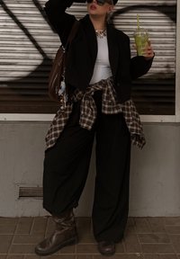 Fekete túlméretezett blézer, fehér testhezálló felső, kockás ing a derékra kötve, széles fekete nadrág, barna térdig érő csizma, zöld italt tartva.