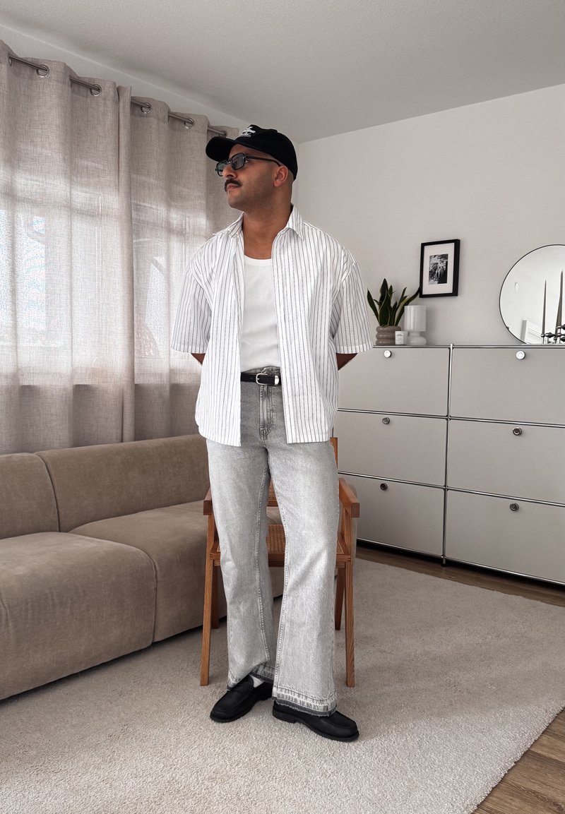 Homme portant une casquette noire, des lunettes de soleil, une chemise blanche à rayures, un débardeur blanc, un jean gris et des chaussures noires, debout dans un salon avec un canapé beige et un meuble blanc.