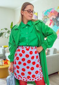 Grønn skjorte med knapper under en lang grønn kåpe, kombinert med et hvitt forkle med røde tomatmønstre og en lys blå håndveske.