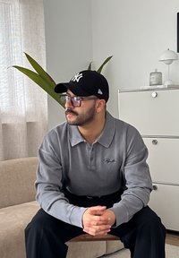 Grå ribbet polo-shirt med krave, båret med sorte bukser. En sort baseball cap og briller komplimenterer antrekket. Indendørs miljø med planter.