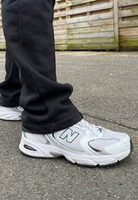 Tenisky New Balance majú vrch z bielej sieťoviny so striebornými akcentmi, hrubú čiernu podrážku a logo značky na boku. Nosené sú čierne teplákové nohavice.