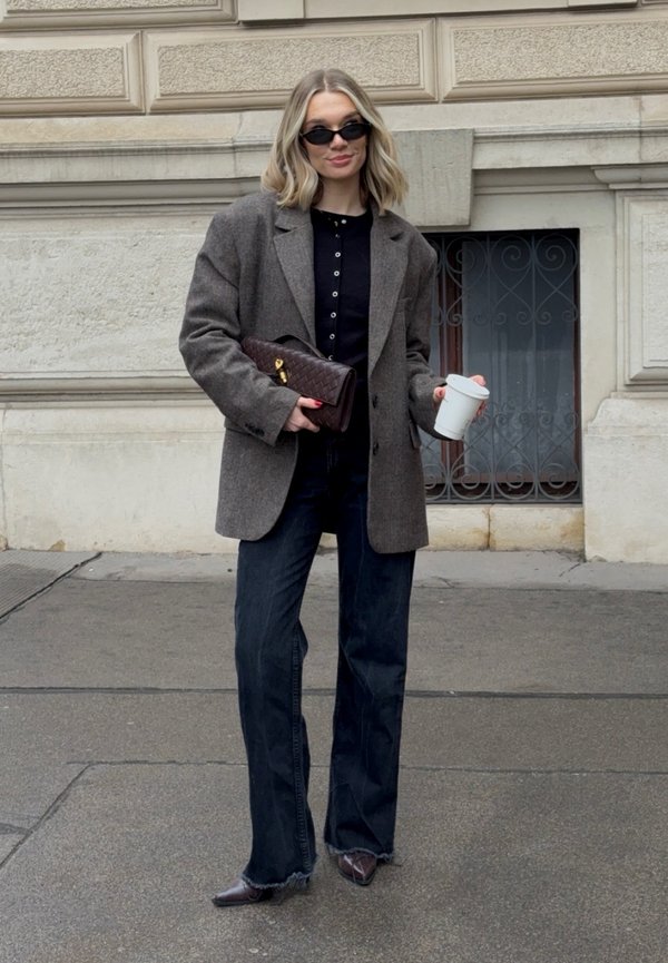 Brauner Blazer aus Wollmischung über einem schwarzen Hemd mit Knöpfen, kombiniert mit dunkelblauen Jeans und spitz zulaufenden braunen Schuhen. Hält eine weiße Tasse und eine gesteppte Handtasche.