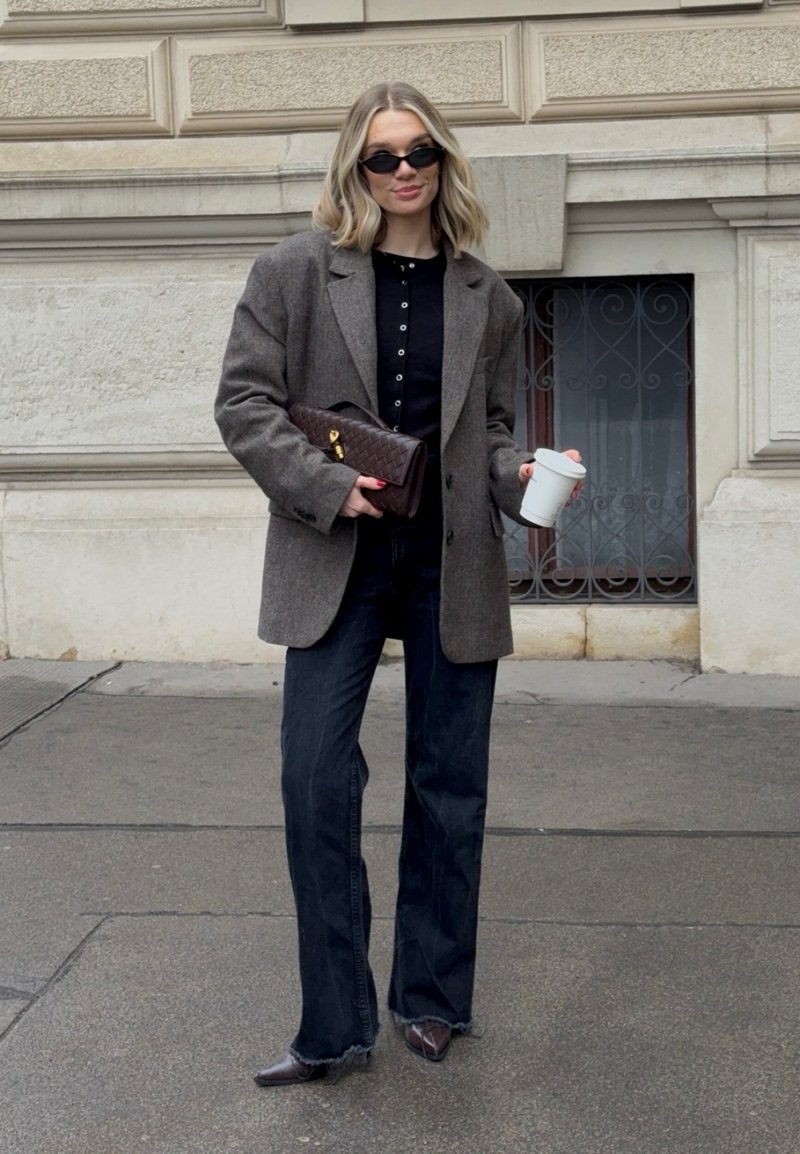 Blazer marron en mélange de laine sur une chemise noire à boutons, associé à un jean en denim foncé et des chaussures marron pointues. Tenant une tasse blanche et un sac à main matelassé.