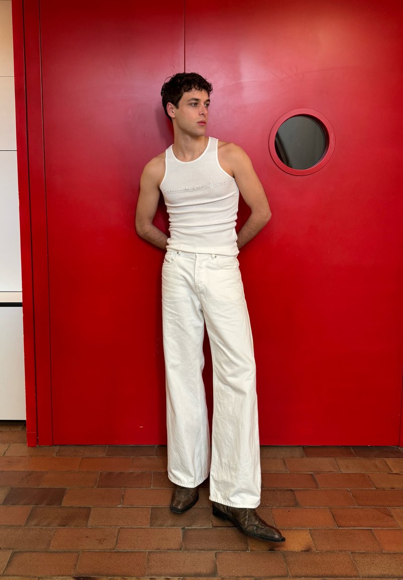 Jeune homme en débardeur blanc et pantalon blanc à jambes larges, debout les mains derrière le dos devant une porte rouge avec une fenêtre ronde.