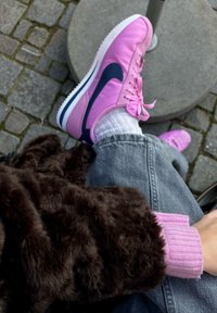 Ténis cor-de-rosa com detalhes em azul marinho, pelo sintético castanho texturizado, punho de suéter em rosa claro com nervuras, jeans cinzentos, posição sentada em calçada de pedras.