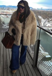 Femme en manteau en fourrure surdimensionné, jean bleu, gants noirs et casquette, debout sur un balcon surplombant une ville enneigée et une rivière.
