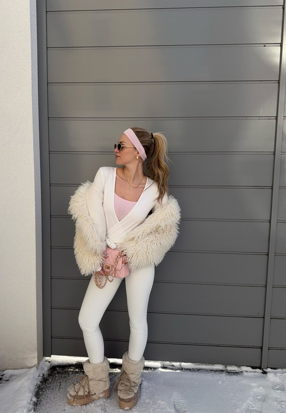 Femme en tenue blanche avec manches duveteuses et bottes, bandeau rose, lunettes de soleil et sac rose, debout sur la neige près d'un mur gris.