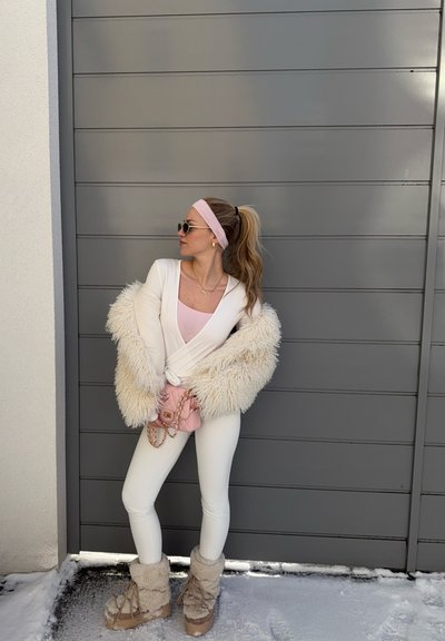 Mujer con conjunto blanco de mangas esponjosas y botas, diadema rosa, gafas de sol y bolso rosa, de pie sobre la nieve junto a una pared gris.