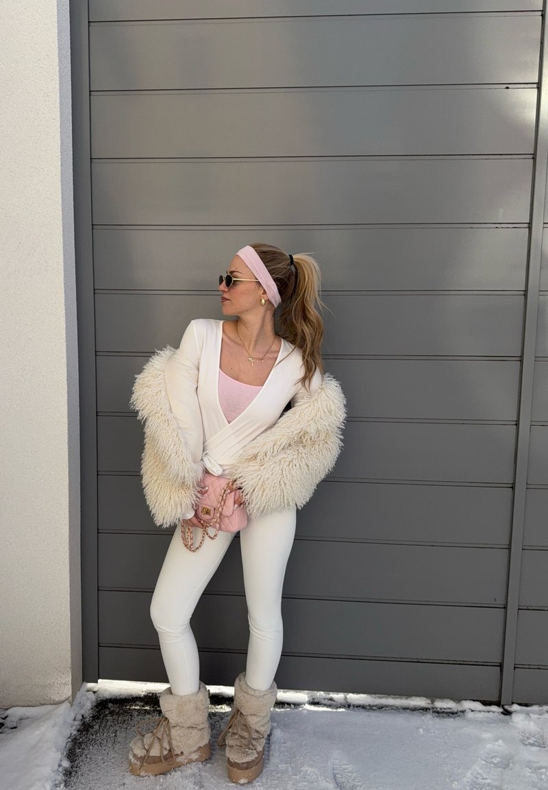Femme en tenue blanche avec manches duveteuses et bottes, bandeau rose, lunettes de soleil et sac rose, debout sur la neige près d'un mur gris.