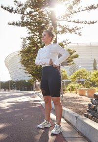 Athletin in weißem Langarmshirt und schwarzen Radshorts, trägt weiße und goldene Sneakers, steht im Freien mit Bäumen und einem Stadion im Hintergrund.