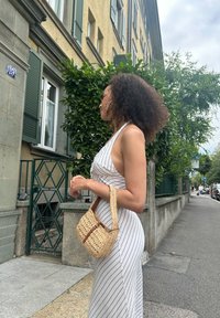 Tkané slamené torbico s kratko ročajem, ki ima črtast vzorec in rjave poudarke, v kombinaciji z oprijeto belo-sivo črtasto obleko.