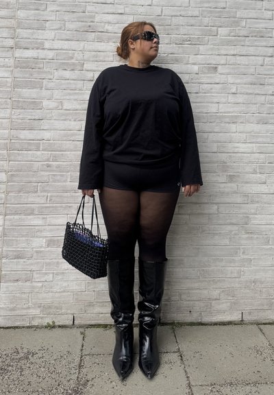 Mujer con top negro de manga larga, pantalones cortos, medias transparentes y botas negras brillantes hasta la rodilla, apoyada contra una pared de ladrillos blanca, sosteniendo un bolso negro tejido.