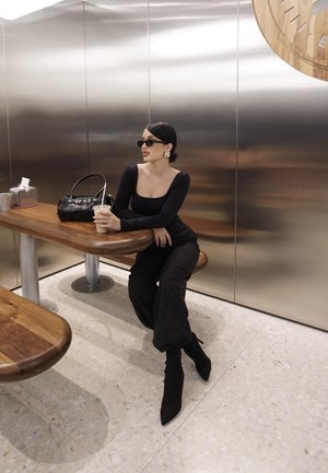 Top negro de manga larga, pantalones negros de tiro alto, botas negras hasta el tobillo. Sentada en una mesa de madera con una bebida, gafas de sol y un bolso negro.