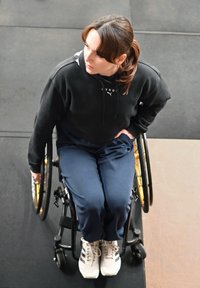 Femme en fauteuil roulant portant un sweat à capuche noir et un pantalon bleu, poussant les roues du fauteuil tout en regardant vers le haut et sur le côté à l'intérieur.