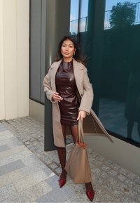 Vestido de couro castanho, casaco bege, mala castanha e saltos altos vermelhos; a de pé sobre paralelepípedos com um fundo de vidro; cabelo longo e solto.