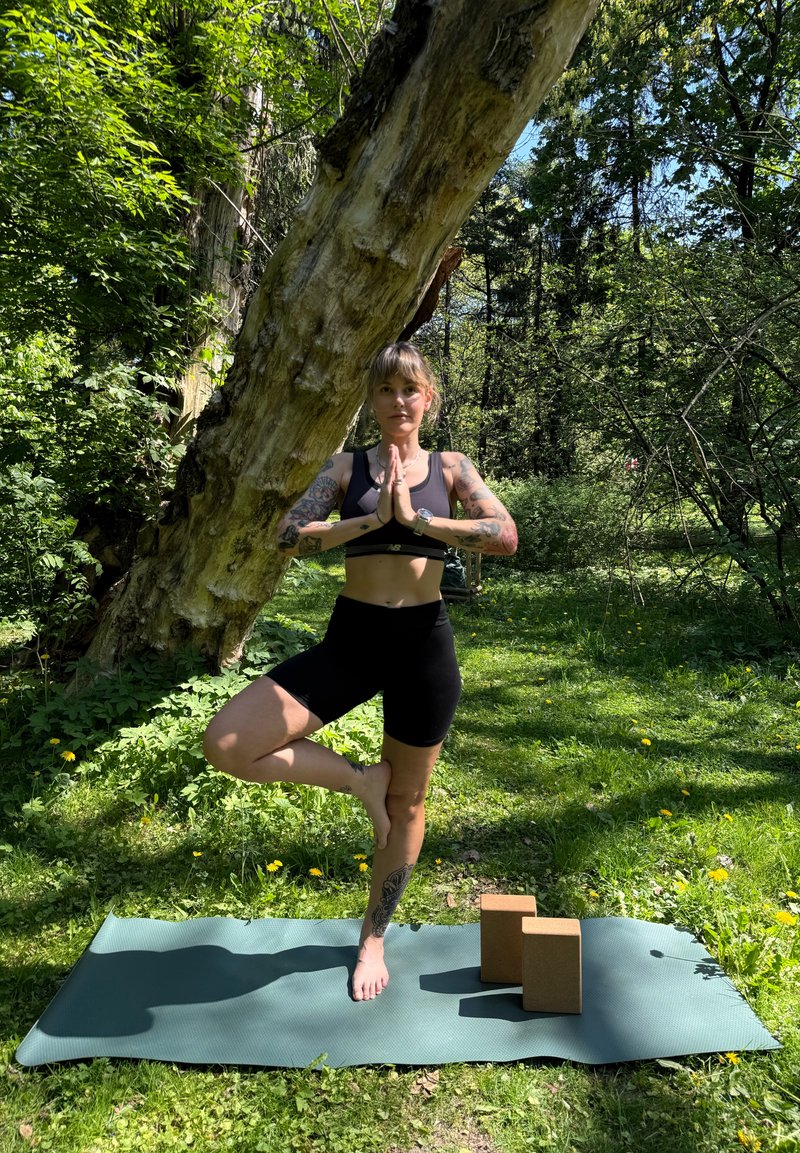 Kvinne med tatoveringer praktiserer trepose yoga på matte utendørs, med to korkblokker i nærheten og et stort tre bak henne.