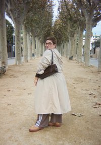 Una persona con capelli corti e occhiali, che indossa un cappotto beige e pantaloni marroni, si trova su un sentiero di terra fiancheggiato da alberi, mentre guarda indietro sopra la spalla.