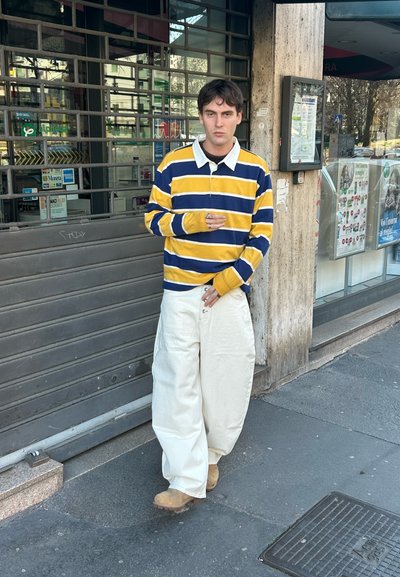 Joven con un suéter a rayas amarillas y azul marino y pantalones blancos anchos, de pie en una acera frente a la persiana cerrada de una tienda.