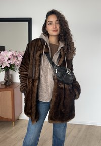 Bruine jas van kunstbont, grijze hoodie, beige trui, blauwe jeans, zwarte heuptas. Heeft krullend haar, staat binnenshuis tussen planten en decoratieve meubels.