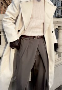 Manteau blanc croisé, pull côtelé crème, pantalon gris plissé et gants en cuir noir. Textures lisses avec une coupe ajustée.
