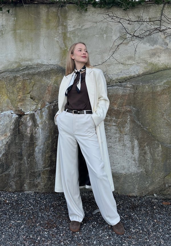 Ženska stoji ob skalni steni, oblečena v belo dolg plašč, črno srajco, bele hlače, črno-belo šal okoli vratu, roke v žepih, gleda navzgor.