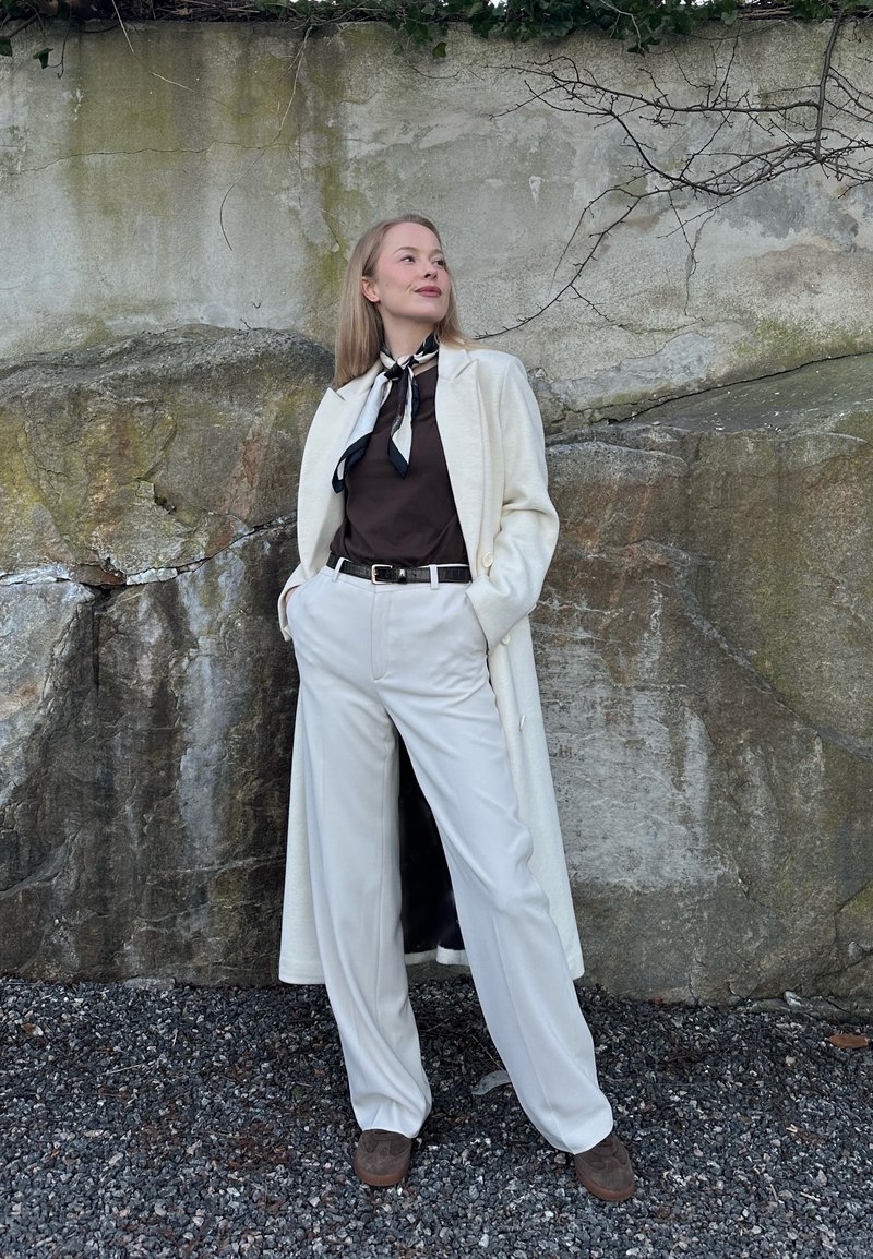 Γυναίκα που στέκεται μπροστά σε βραχώδη τοίχο, φοράει λευκό μακρύ παλτό, μαύρο πουκάμισο, λευκό παντελόνι, ασπρόμαυρο φουλάρι στο λαιμό, τα χέρια στις τσέπες, κοιτάζει προς τα πάνω.