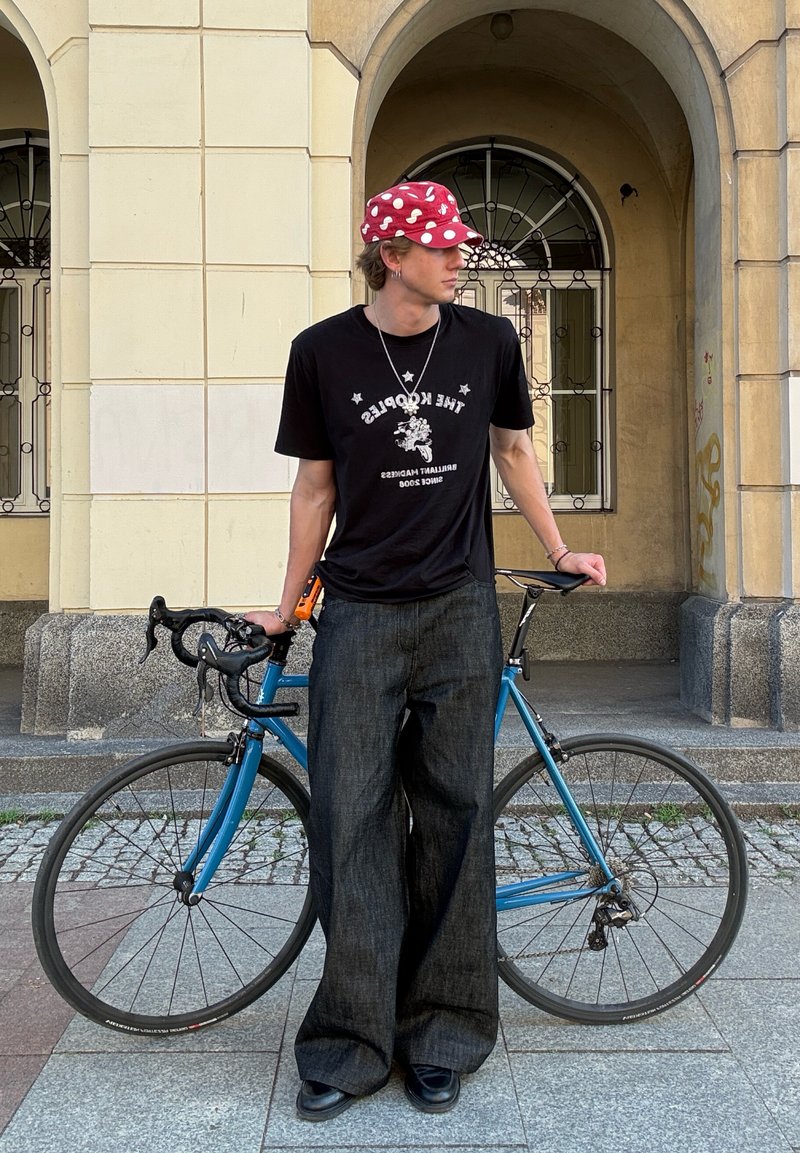 Jonge man in een zwart grafisch t-shirt, wijde jeans en een rode stippenpet staat met een blauwe racefiets voor gebogen stenen ramen.