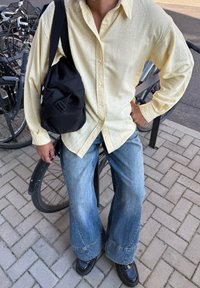 Világossárga, hosszú ujjú, laza fit szabású ing; fakó kék, bő szárú farmernadrág; fekete kereszttestű táska; fényes fekete cipők, kerékpárok mellett állva.