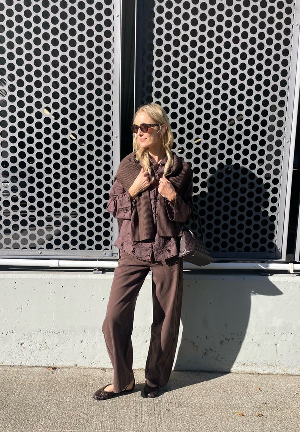 Brun lagd outfit med en tekstureret overdel, matchende bukser og sjal. Accessories inkluderer solbriller og en mønstret håndtaske. Baggrund med polka prikker.
