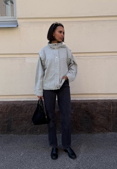 Camisa gris, texturizada, con rayas horizontales, cierre de botones y cuello embudo. Combinada con pantalones oscuros de pierna recta y un bolso negro.