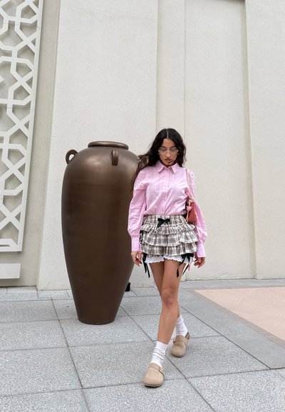Mujer con camisa rosa y falda de cuadros en capas caminando junto a un gran ánfora marrón en un patio al aire libre con suelo de baldosas.