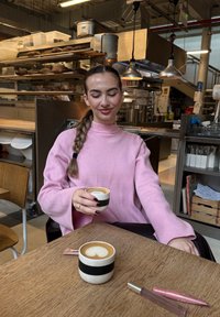 Sweater in soft pink fabric with wide sleeves, holding a coffee cup with a cream stripe. Wooden table with a second cup and utensils visible.