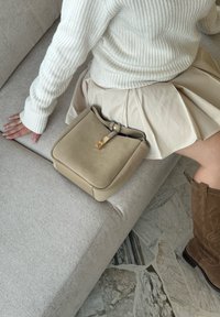 Beige mokkanahkakäsilaukku, jossa on jämäkkä muoto, kultaiset yksityiskohdat ja säädettävä olkahihna. Aseteltu vaaleanharmaalle sohvalle kermaisesti värjätyn hameen viereen.