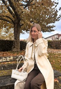 Casaco trench de creme com gola larga e botões, combinado com calças castanhas. Uma mala branca texturizada com ferragens douradas está ao lado dela num banco.