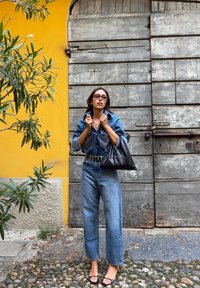 Traper košulja i traperice visokog struka, oboje svijetloplave boje. Crni remen, prevelika crna torba i sandale na remenčiće. Drvena pozadina.
