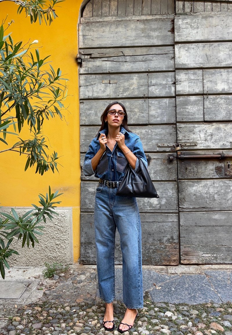 Camisa de denim e jeans de cintura alta, ambos em azul claro. Cinto preto, mala oversized preta e sandálias com tiras. Fundo de madeira.