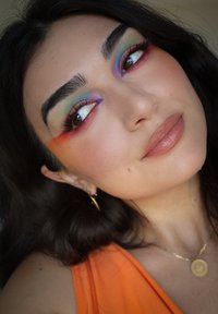 Närbild av ett ansikte med färgglad ögonmakeup i gröna, lila och röda nyanser. Slät hy, ljusbruna läppar och gyllene ringörhängen.