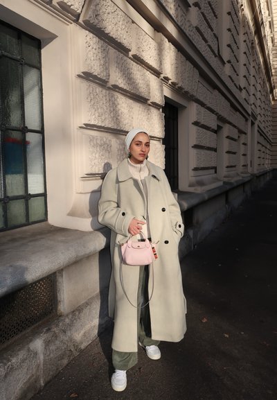 Abrigo de lana verde claro con botones negros, combinado con un suéter blanco y pantalones verdes anchos. Pequeño bolso rosa y zapatillas blancas.