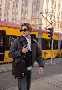 Pilkas korduro pluošto švarkas dėvimas ant tamsiai mėlynos džemperio su gobtuvu, derinamas su mėlynais džinsais. Komplektą sudaro tamsios medžiagos peties krepšys su matomomis metalinėmis detalėmis.