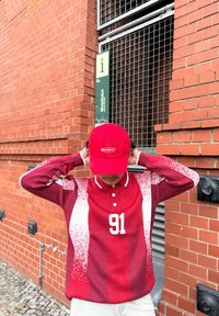 Rotes langärmeliges Polohemd mit weißen Akzenten und einer großen "91" auf der Vorderseite, kombiniert mit einer passenden roten Kappe mit einem Logo.