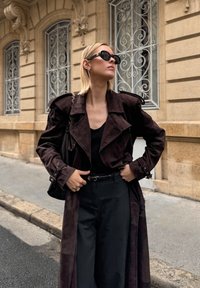 Σκούρο καφέ σουέτ παλτό τέντζερ με μεγάλα πέτα, μαύρη μπλούζα, ψηλόμεσα μαύρα παντελόνια και στρογγυλά μαύρα γυαλιά ηλίου, μπροστά από έναν πέτρινο τοίχο.